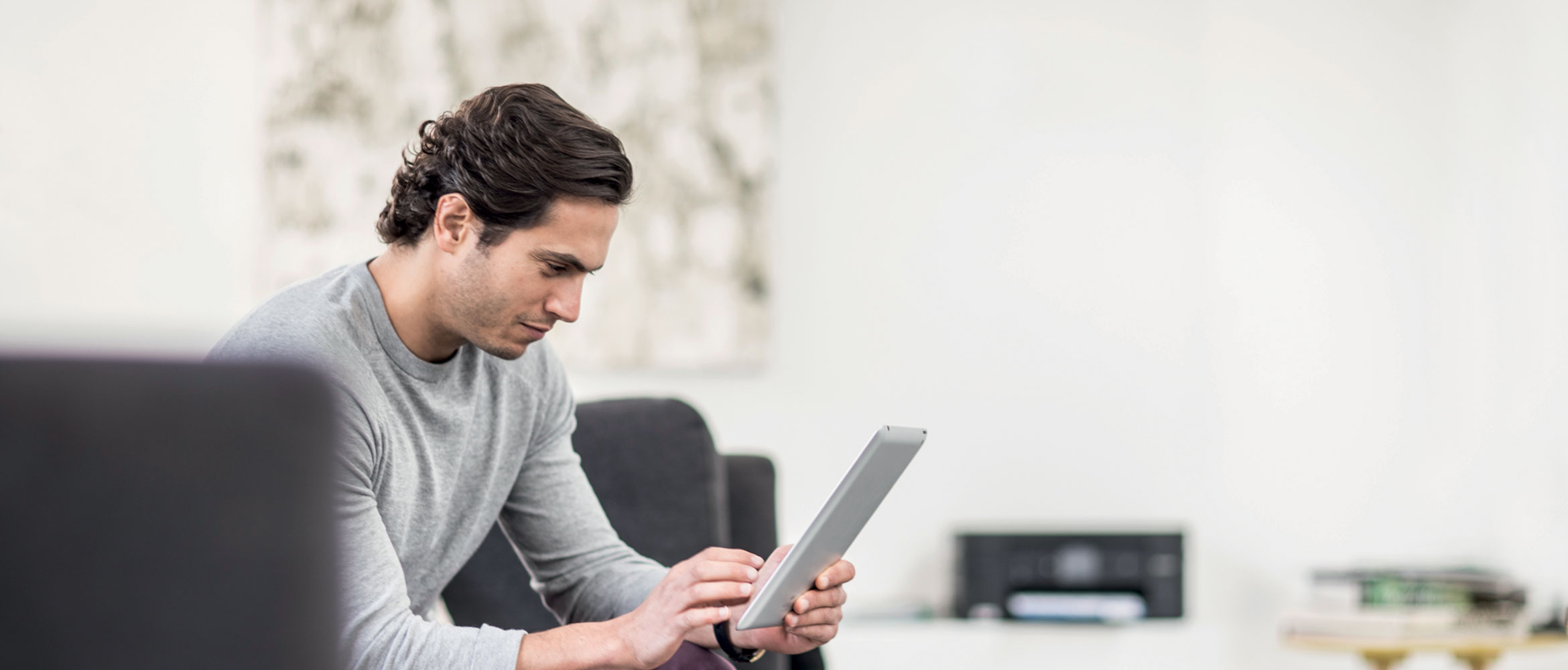 Man looking at tablet sat on sofa in home, with Brother printer in the background