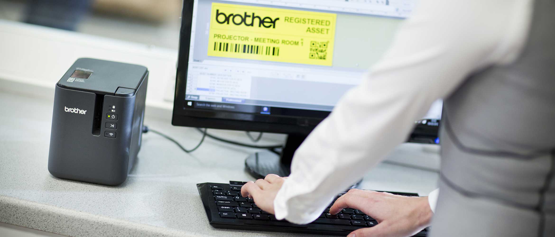 Drukarka etykiet Brother PT-P900 stoi na biurku obok monitora 