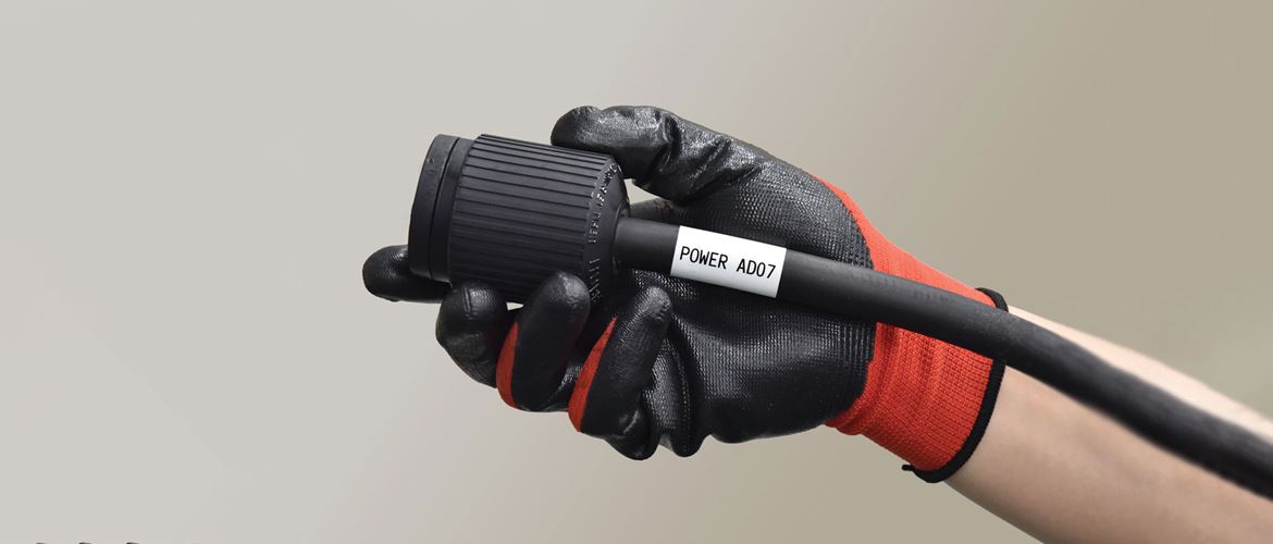 Brother Pro Tape flexible ID label on a thick industrial electrical cable, being held by a construction worker in a gloved hand