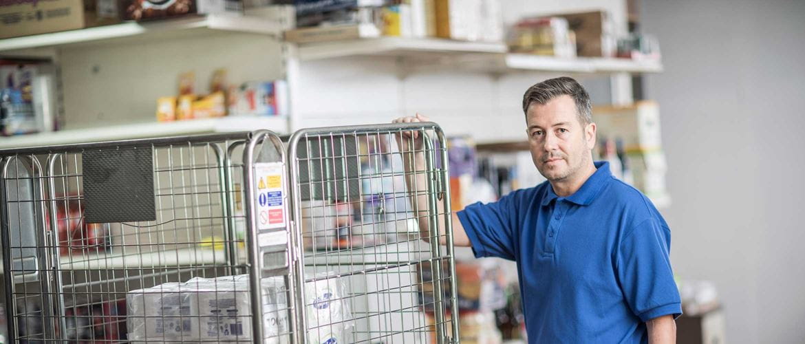 mężczyzna w niebieskiej koszulce polo stoi na zapleczu w tle półki z produktami 