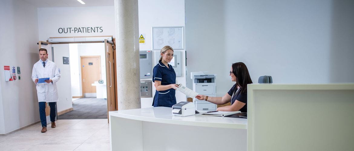 Receptionist wearing glasses sat at desk with nurse and doctor working in background