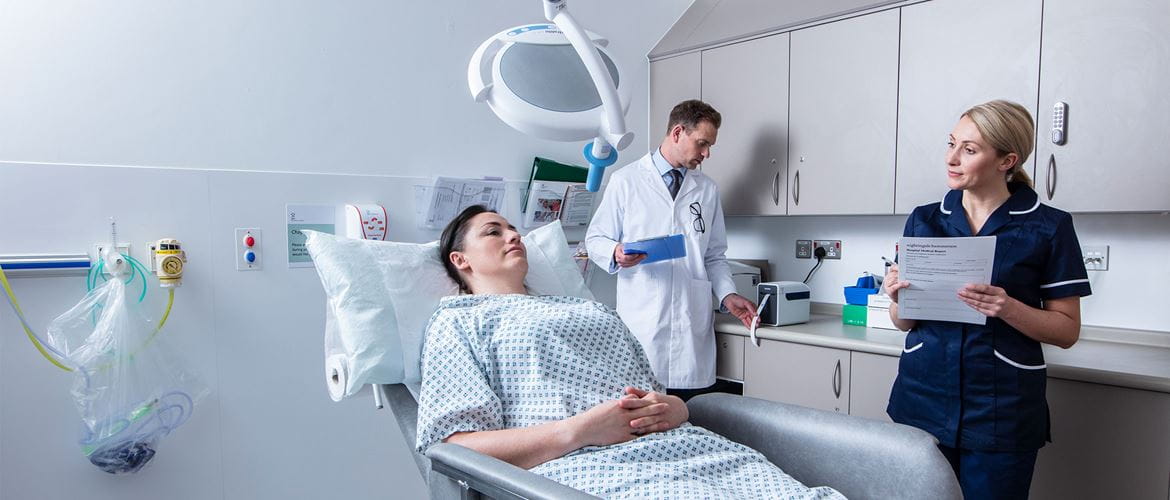 Pacientka ležiaca v nemocničnej izbe s lekárom v bielom kabáte, vzadu stojaca zdravotná sestra s formulárom