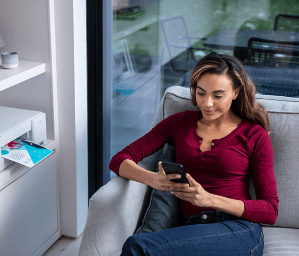 Nainen istuu sohvalla tulostin hyllyssä vieressään ja selaa älypuhelintaan