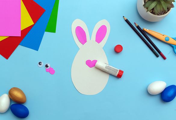 Kuvassa erivärisiä pääsiäismunia, kyniä, sakset ja sininen korttipohja, jossa on pääsiäispupu
