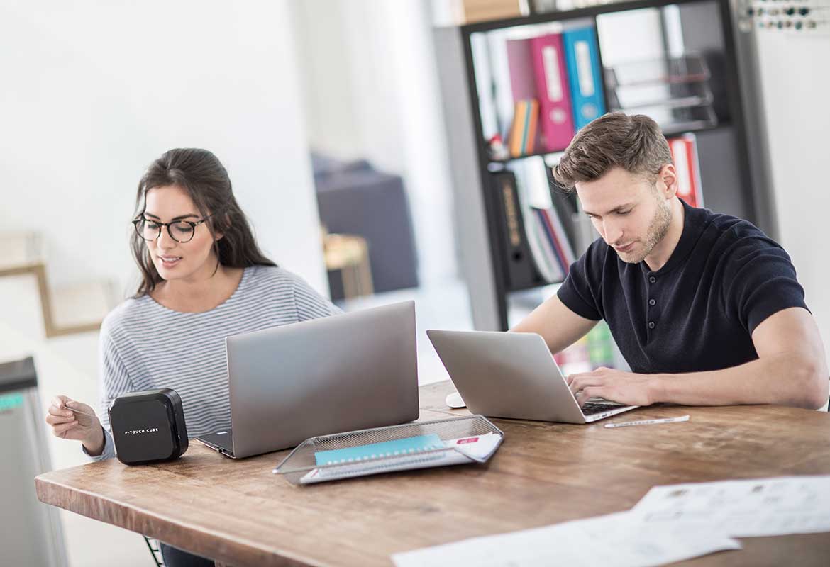 A man and a woman in a smart modern office use their computers to design and print labels to a Brother P-touch label printer