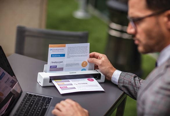 Male sat at grey table wearing glasses, grey suit with laptop scaniing colour document outdoors 