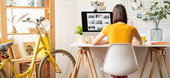 žena zu žutoj majici okrenuta leđima piše na računalo u bijelom uredu i žutom biciklom