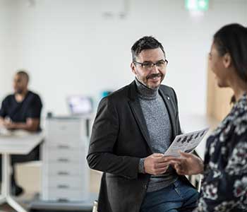 Male sat in office holding document speaking to female, people and printer in background