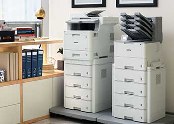 Two floor standing printers side by side against a wall, cabinets, folders, picture frames