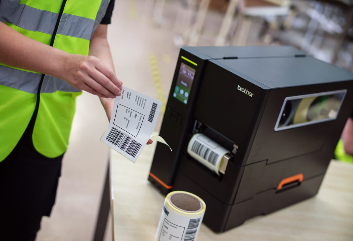 En lagerarbeider med synlig gul vest som tar av baksiden av en utskrevet etikett printet fra en Brother industriell etikettskriver og rekvisita på et bord 