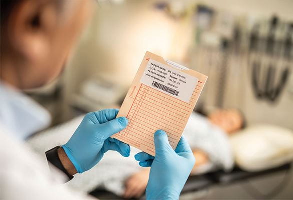 Férfi orvos kék gumikesztyűt visel, tart egy betegkartont, a beteg adataival a címkén, női beteg fekszik az ágyon, orvosi berendezésekkel a háttérben