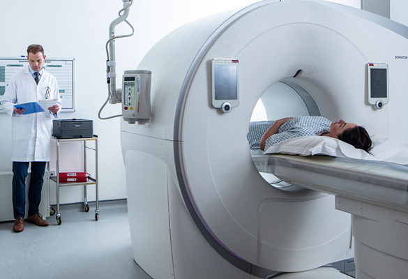 Pacientka v CT-skenerju, zdravnik v uniformi drži modro mapo s sponko in stoji poleg tiskalnika Brother HL-L2370DW