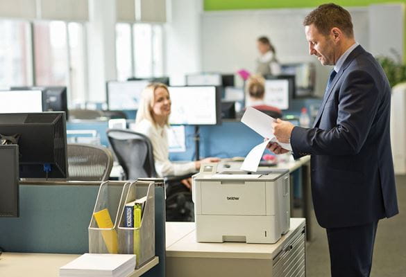 Man wearing suit standing at printer holding paper, monitors, woman, office, table