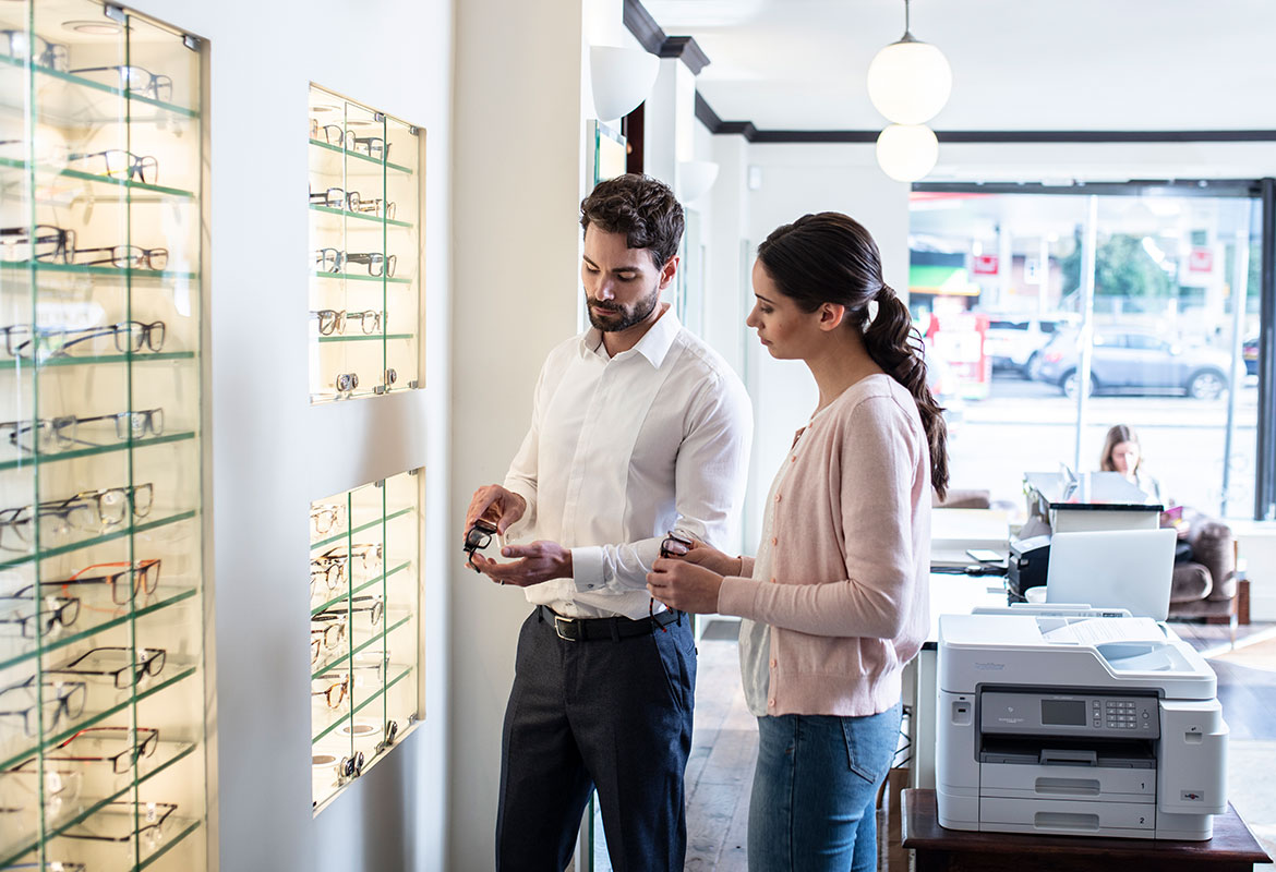 Man in white shirt holding glasses, woman with long brown hair in pony tail wearing pink cardigan, jeans, Brother printer, glasses, laptop