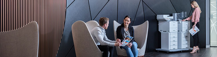 Woman stood at two floor standing printers side by side, man, woman sat on chairs