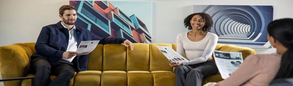 Mies ja nainen istuvat sohvalla esitteet kädessään ja etualalla istuu nainen selin kuvaajaan