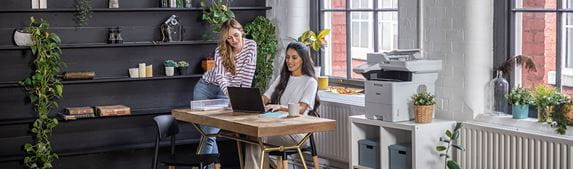 Two women sat around desk in small business, plants, Brother printer, cabinet