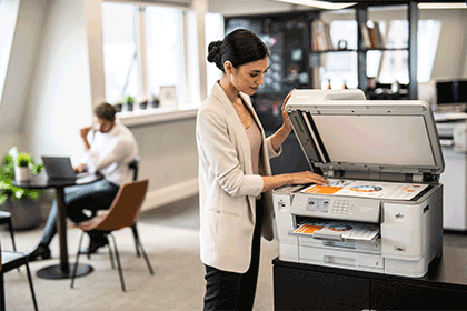Woman in office scanning document in printer, man in background, chair, table, plants