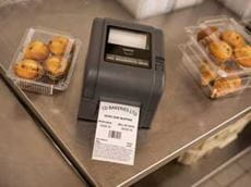 Drukarka etykiet Brother TD-4D drukuje etykietę stojąc na metalowym blacie w towarzystwie ciastek