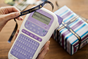 Someone using a P-touch handheld label printer to print "Happy Birthday" in gold lettering onto a black ribbon to tie around a wrapped gift