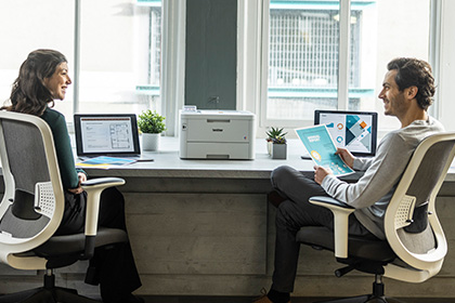 Female and male sat at desk, laptops, Brother HL-L8240CDW printer, window, chairs, plants