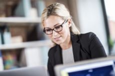 En kvinne i sort dressjakke og briller sitter på et kontor å jobber på datamaskinen