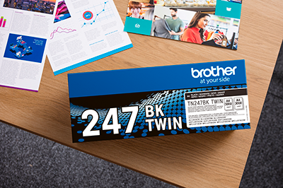 En eske med Brother original TN247BKTWIN sort toner i Twin Pack ligger på et bord sammen med fargerike dokumenter