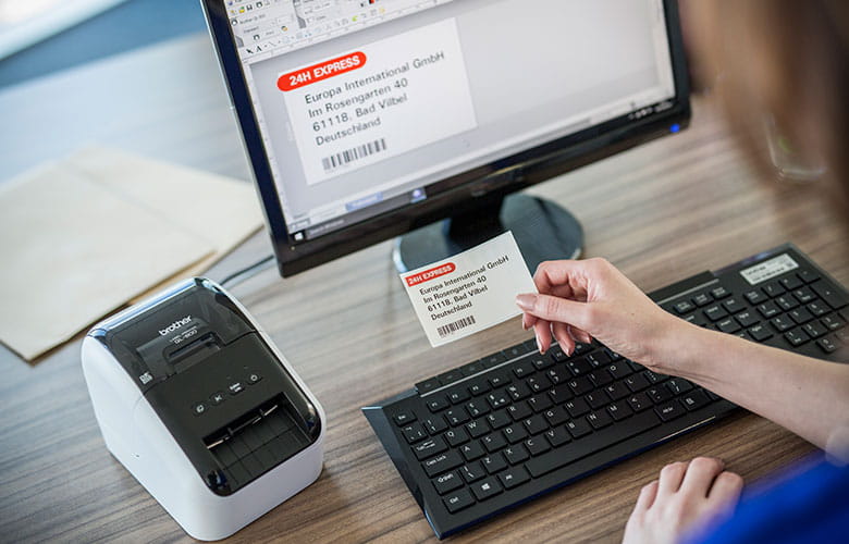 En person holder en utskrevet etikett som er skrevet ut på en Brother QL etikettskriver som er plassert på et skrivebord ved siden av en dataskjerm og tastatur