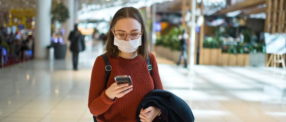 Une jeune femme se promenant dans un centre commercial consulte son smartphone tout en portant un masque. 