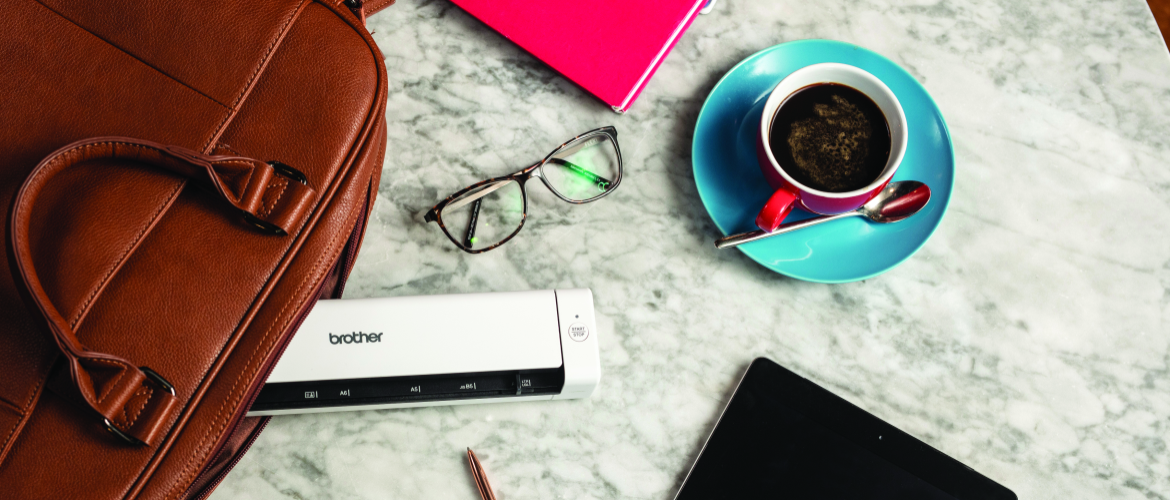 Plusieurs objets sont posés sur une table en marbre, parmis eux des lunettes de vue, une tasse à café turquoise et une sacoche de laquelle dépasse un scanner portable Brother.