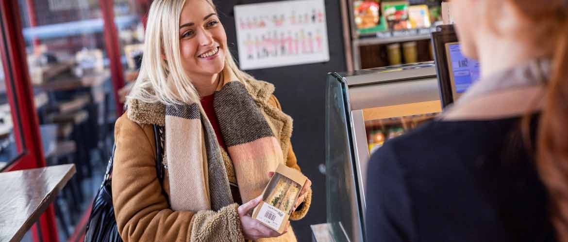 Une jeune femme aux cheveux blonds, vêtue d'un manteau et d'un foulard d'automne, achète un sandwich pré-préparé et clairement étiqueté dans une épicerie de détail. Le client est servi par une employée du magasin derrière la caisse.