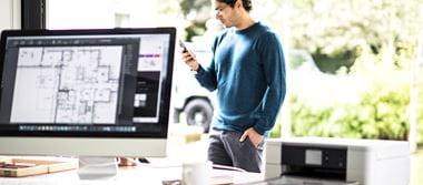Un employé regarde son smartphone. Au premier plan, son bureau équipé d'un ordinateur et d'une imprimante Brother