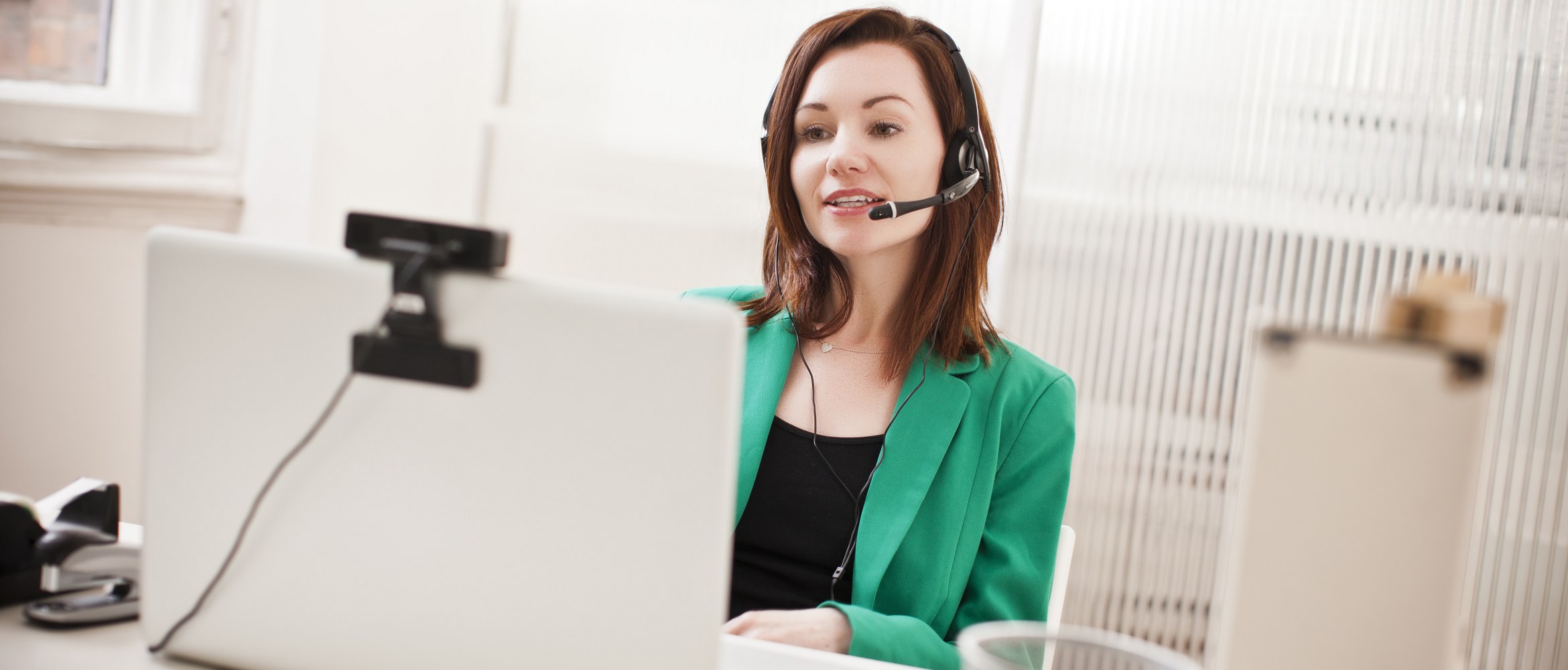 Une femme portant un micro-casque et une veste verte participe à une visioconférence au sein d'un petit bureau à domicile. 