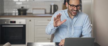 Un homme en télétravail dans sa cuisine salue un collègue d'un geste de la main via son ordinateur