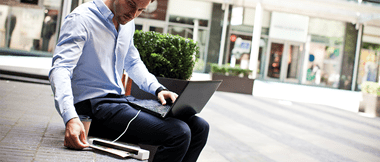 Un homme assis en extérieur tient un ordinateur sur ses genoux et utilise un scanner portable pour numériser un document. 