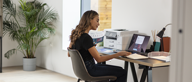 Une femme est en train de travailler depuis sa maison. Elle est assise devant son bureau.  La femme travaille sur son ordinateur, à côté d'elle se trouve une imprimante. En fond, une plante verte est visible. 