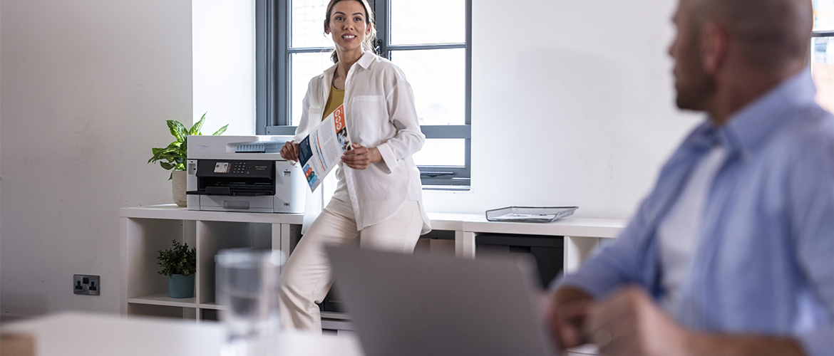 Un homme assis à un bureau regarde derrière lui une femme tenant des documents dans sa main. Derrière elle on peut voir un imprimante.