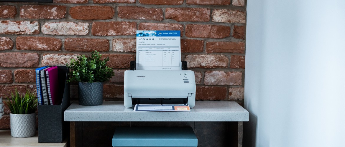 Un scanner Brother placé au-dessus d'un bureau, devant un mur de briques apparentes, avec un papier imprimé dans le bac de sortie