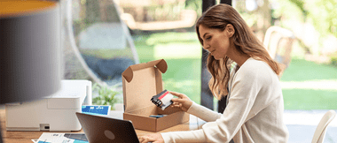 Une femme assise devant son ordinateur tient dans sa main droite une cartouche d'encre pour imprimante