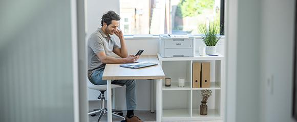 Homme assis sur une chaise à un bureau regardant son téléphone près d'une fenêtre avec une imprimante, des dossiers et des plantes sur des étagères.