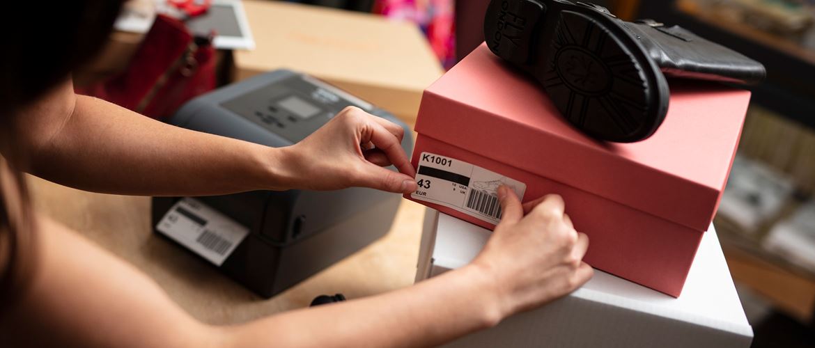 Une femme colle une étiquette de boîte à chaussures sur une boîte à chaussures rose devant une imprimante d'étiquettes de bureau Brother TD.