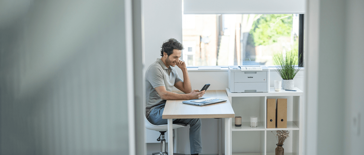 Homme assis sur une chaise à un bureau regardant son téléphone près d'une fenêtre avec une imprimante, des dossiers et des plantes sur des étagères.