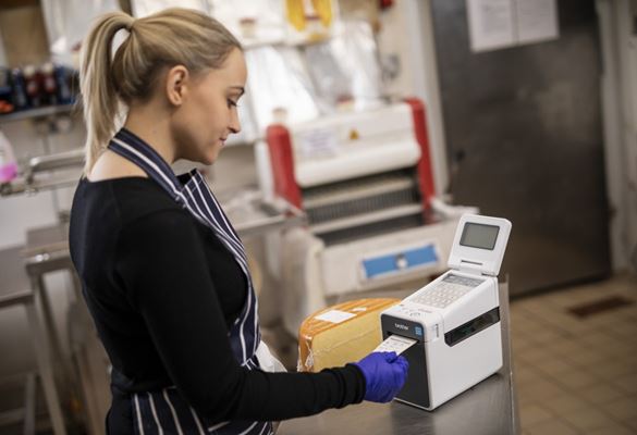 Une employé imprime une étiquette alimentaire à partir d'une étiqueteuse Brother pour l'ajouter à une meule de fromage dans l'arrière-boutique d'un traiteur ou d'un café. 