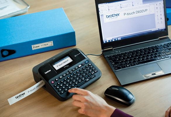 Imprimante d'étiquettes P-touch posée sur un bureau, connectée à un ordinateur portable, en train d'imprimer une étiquette.