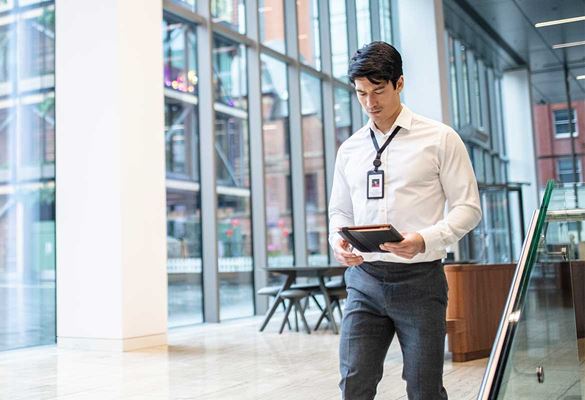Homme en chemise portant un badge de bureau et un livre marchant dans une entreprise avec des murs en verre.