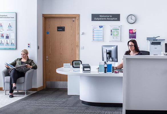 Réceptionniste assis à son bureau regarde son ordinateur et une patiente assise lisant un journal