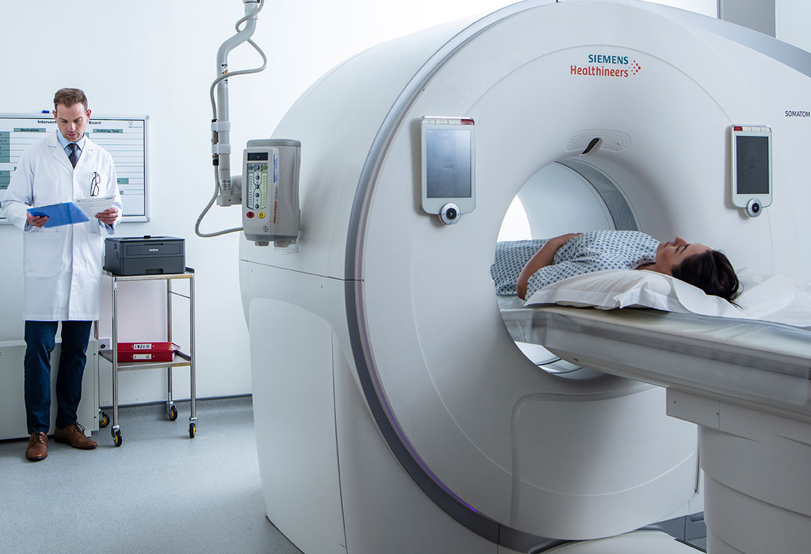 Une patiente dans un scanner médical et médecin tenant un presse-papiers bleu avec une imprimante Brother HL-L2370DW posée à côté