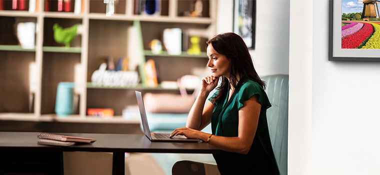 Woman with shoulder length brown hair in green sat using laptop at table, books, picture on wall, vase, ornaments