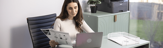 Un femme assise à son bureau travaille sur son ordinateur et tient dans son autre main des documents.