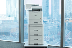 Imprimante multifonction posée sur un plateau avec plusieurs bacs de papier dans un bureau, grands bâtiments, grandes fenêtres
